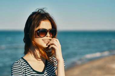 Tişört ve gözlüklü bir kadın kumlu sahilde mavi denizin yanında yürüyor.