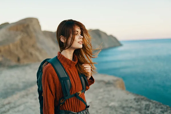 Viaggiatore felice in un maglione rosso con uno zaino sulla schiena guarda il mare in lontananza e in alta montagna — Foto Stock