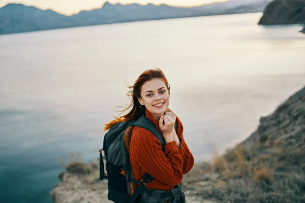 Excursionista con una mochila en las montañas cerca del mar y un suéter rojo pelo suelto puesta de sol rocas — Foto de Stock