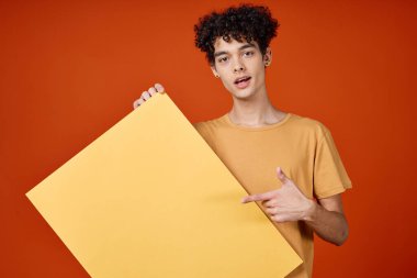 Kıvırcık saçlı, neşeli bir adam. Stüdyo reklamında sarı afiş asmış.