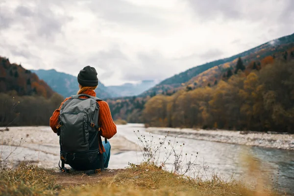 一个女人，背着背包，穿着一件夹克，戴着一顶帽子，站在山中的河岸上 — 图库照片