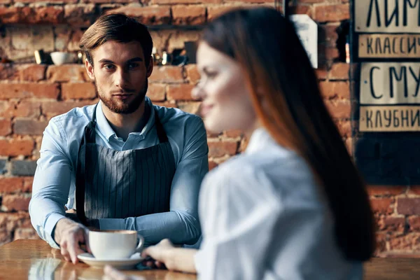 Mujer feliz con una taza de café y camarero masculino en el delantal en la mesa en el interior — Foto de Stock