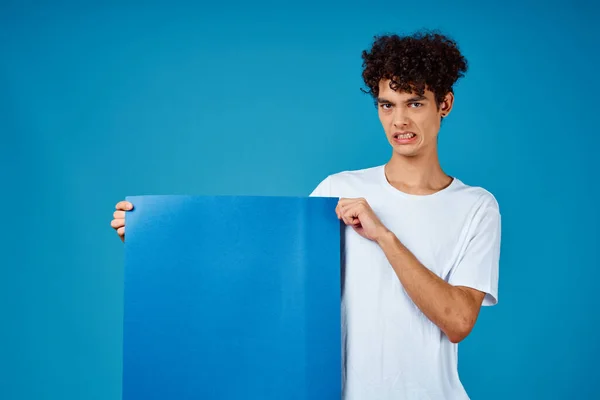 Cara em um branco t-shirt segurando um cartaz mockup publicidade azul fundo — Fotografia de Stock