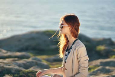 Gözlüklü bir kadın ve doğa manzarasında deniz kenarındaki dağlarda süveter giyen bir kadın.