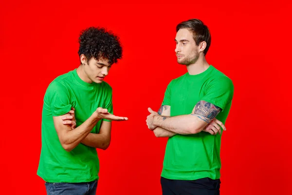 녹색 티셔츠를 입은 두 남자가 의사소통하는 우정의 붉은 배경 옆에 서 있다 — 스톡 사진