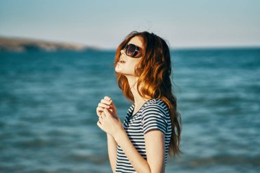 Tişört giyen, deniz kenarında güneş gözlüklü ve arka planda dağlar olan bir gezgin.