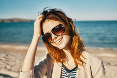 Gözlüklü, bej ceketli, gözlüklü, şık bir kadın. Dağlarda, deniz kıyısında, yakın plan bir portre.