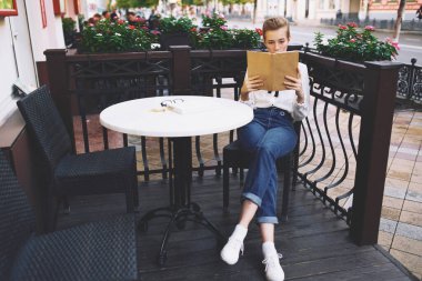 Elinde kitap olan şık, genç bir kadın yazın bir kafede bir masada oturuyor.