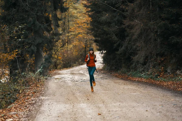 Viaje feliz con una mochila en un suéter cálido, pantalones y botas camina a lo largo de la carretera en el bosque de otoño — Foto de Stock