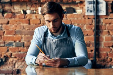 Önlüklü bir adam masada oturur ve bir garsonun siparişini kabul eder.