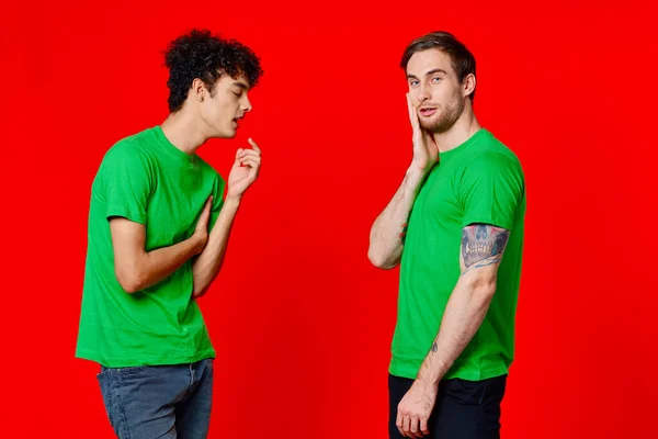 녹색 티셔츠를 입은 두 남자가 의사소통하는 우정의 붉은 배경 옆에 서 있다 — 스톡 사진