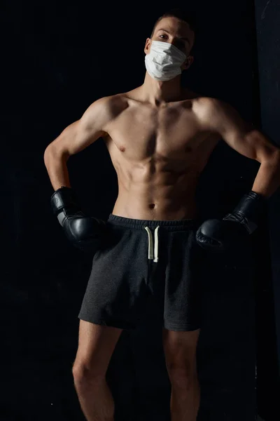 Hombre deportivo en máscara médica y en guantes de boxeo en pantalones cortos de fondo negro fitness — Foto de Stock