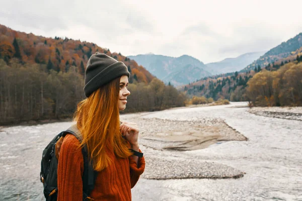 Mulher ruiva em uma camisola com uma mochila e em um chapéu perto de um rio de montanha na natureza — Fotografia de Stock
