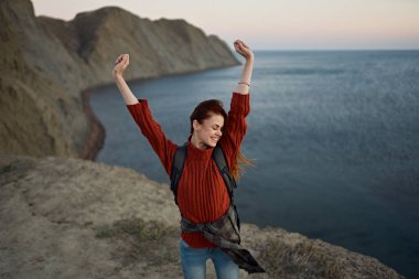 Kollarını kaldırmış mutlu bir kadın deniz kenarında sahilde yürüyor ve yüksek dağlar en tepede geziniyor.