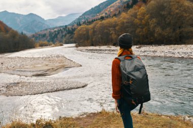 Kırmızı ceketli ve sırtında sırt çantası olan bir kadın dağlardaki nehrin kıyısında yürüyor.