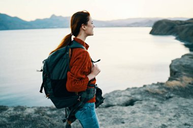 Kadın turist gün batımında deniz kenarında sırtında bir sırt çantasıyla dağlarda