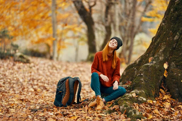 Kobieta w spodniach i butach z plecakiem siedzi w pobliżu drzewa w jesiennym lesie spadły liście — Zdjęcie stockowe