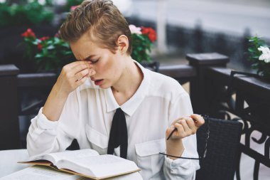 Restoranda bir masada kitap okuyan bir kadın eğitim duyguları arka planda çiçekler gözlüyor.