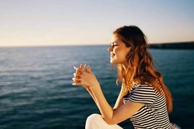 Çizgili tişörtlü ve beyaz pantolonlu bir kadın yazın deniz kenarında bir taşın üzerinde oturur.