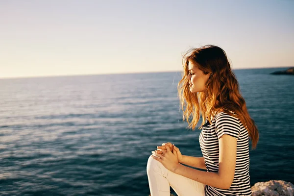 A woman in a striped T-shirt and white trousers sits on a stone near the sea in summer — Stock Photo, Image