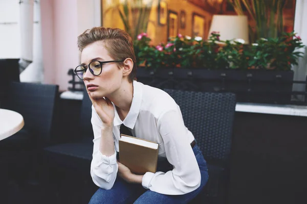 Gebildete Frau mit Buch und Brille sitzt auf Sessel im Café — Stockfoto