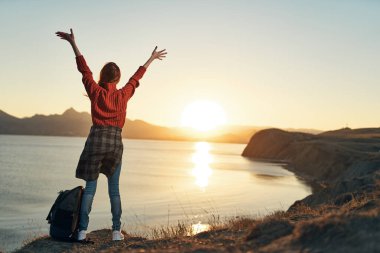 Elleri havada neşeli bir kadın gün batımına bakıyor. Seyahat özgürlüğü.