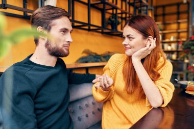 Bir adam, bir kafede bir masada, süveter giymiş genç bir kadına bir şeyler anlatıyor.