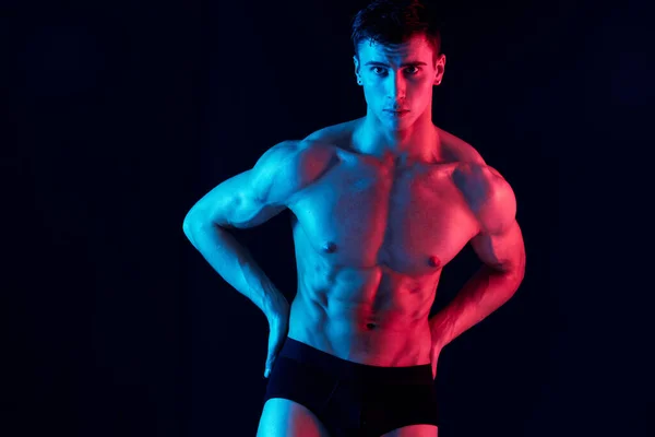 Sexy hombre atleta en pantalones cortos toma las manos en su cinturón en un fondo negro y luz de neón —  Fotos de Stock