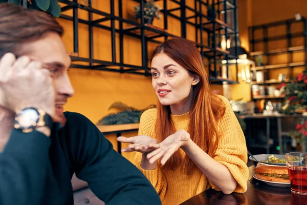 Süveter giymiş mutlu bir kadın elleriyle jest yapar. Duyguları eğlencelidir ve kazaklı bir adam kafede oturur. — Stok fotoğraf