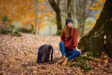 Sonbaharda sıcak giysiler içinde bir kadın ormanda bir ağacın yanında oturuyor.