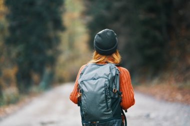 Sırt çantalı, şapkalı ve turuncu kazaklı bir kadın sonbahar ormanı manzaralı yolda.
