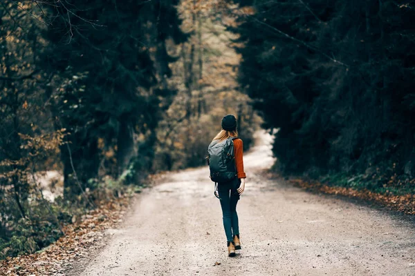 Mulher de casaco jeans com uma mochila caminha ao longo da estrada na floresta de outono — Fotografia de Stock