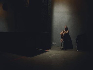 lonely woman sitting on a chair in a dark room leaning against a wall in full growth 