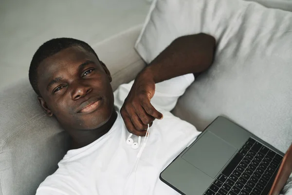Африканський чоловік з ноутбуком лежить на дивані фрілансер, що працює за межами офісу — стокове фото