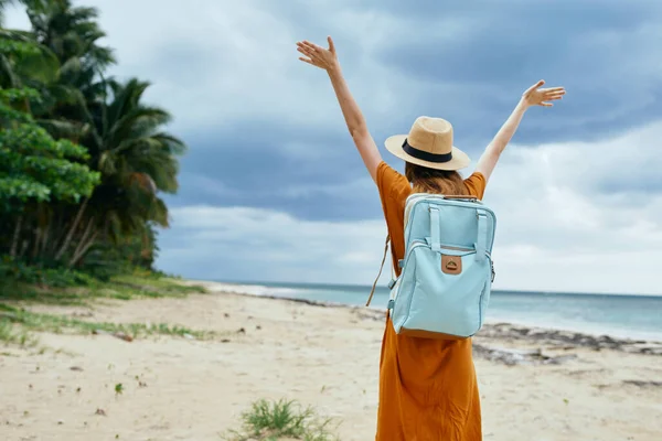 Una mujer con una mochila azul y con un vestido levantó las manos en la isla cerca del mar —  Fotos de Stock