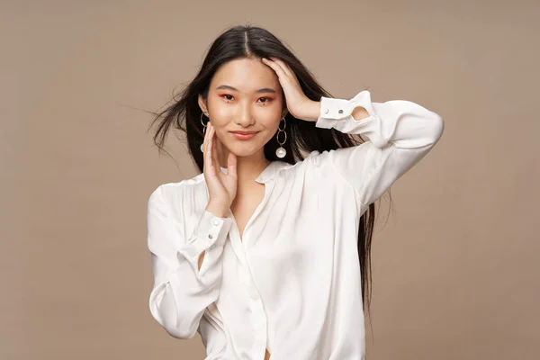 Schöne asiatische Frau in einem hellen Hemd berührt ihren Kopf und lächelt auf einem beigen Hintergrund — Stockfoto