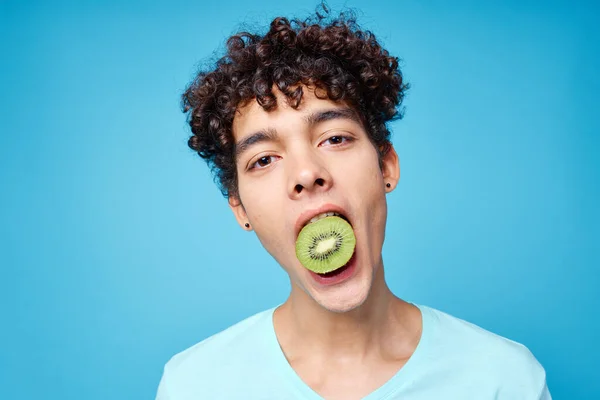 Hombre guapo con el pelo rizado kiwi en su boca vista recortada en fondo azul — Foto de Stock