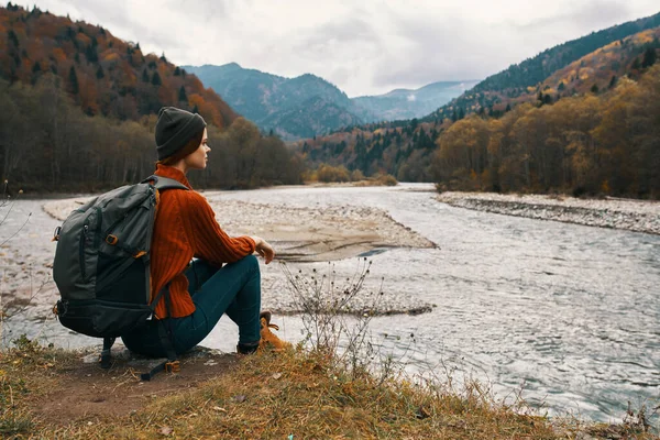 Femme avec un sac à dos dans une veste et un chapeau sur la rive de la rivière dans la vue sur les montagnes — Photo