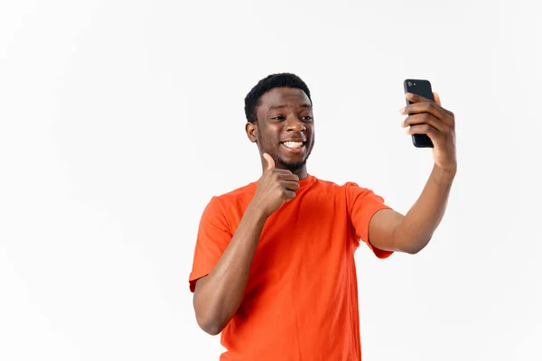 Homem africano feliz em camiseta laranja com telefone celular no fundo claro — Fotografia de Stock