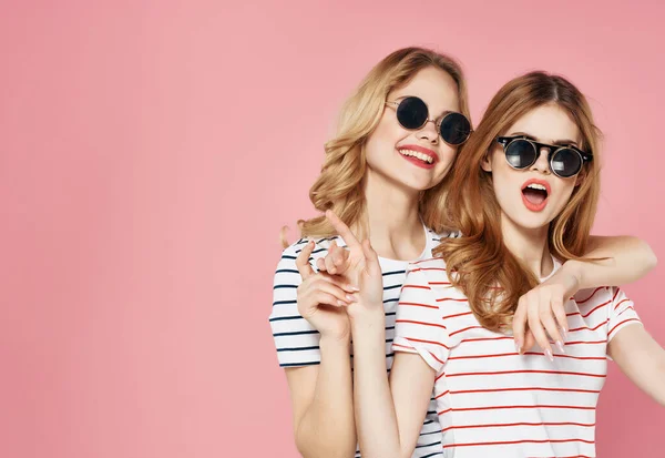 Két barátnő napszemüvegben csíkos pólóban rózsaszín háttér kommunikáció — Stock Fotó