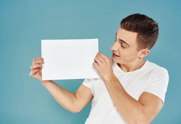 Knappe man in wit t-shirt met wit vel papier in zijn handen paperspace blauwe achtergrond — Stockfoto