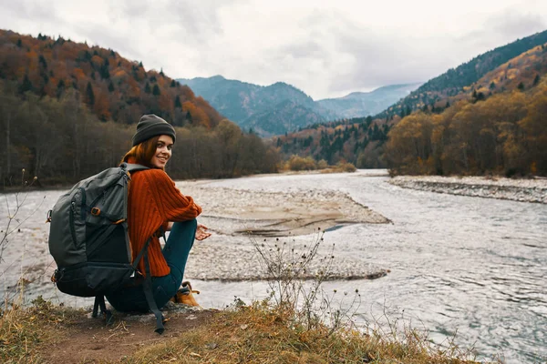 Жінка в горах восени з рюкзаком сидить на березі річки і дивиться на високі гори — стокове фото