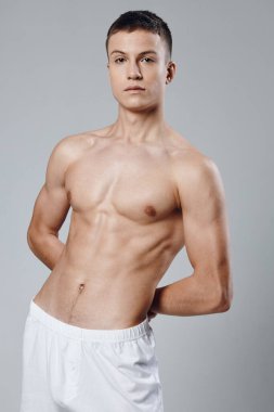 Gri arka planda şişmiş bir gövdesi olan sporcu ellerini arkada ve beyaz şortuyla tutuyor.