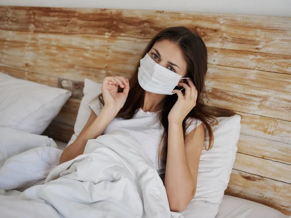 Женщина в медицинской маске лежит в постели под одеялом и жестикулирует руками — стоковое фото