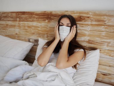 Tıbbi maskeli bir kadın yastık salgınında yatakta yatıyor. Coronavirus akciğer hastalığı karantinasında.