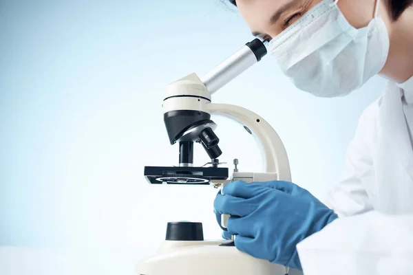 Asistente de laboratorio en una máscara médica mirando a través de un microscopio —  Fotos de Stock