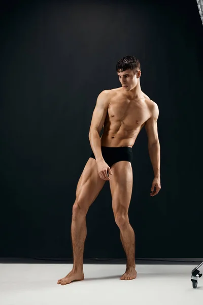 Man with a pumped up naked body in black panties posing against a dark background — Stock Photo, Image