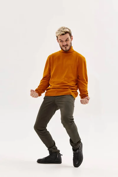Счастливый мужчина в оранжевом свитере и брюках жестикулирует руками на светлом фоне и согнул колени — стоковое фото