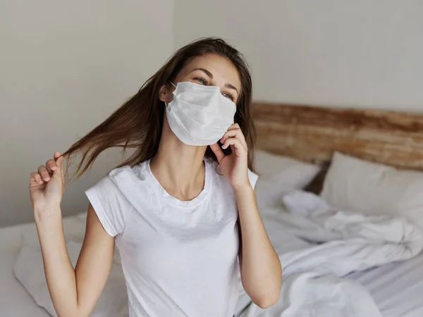 Junge Frau in medizinischer Maske berührt Haare mit der Hand und sitzt drinnen im Bett — Stockfoto