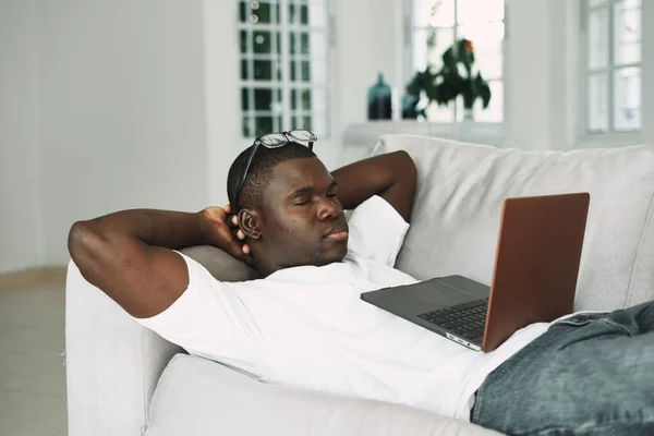 Человек африканского внешнего вида с ноутбуком лежит на диване отдыха — стоковое фото